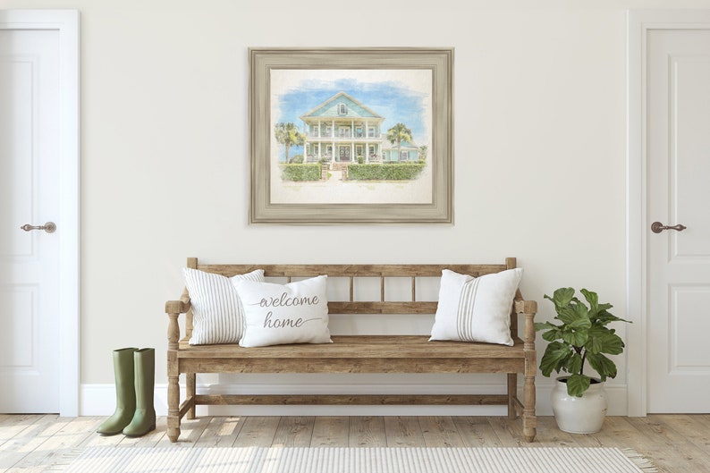 May include: A watercolor painting of a light blue house with white trim and a white porch. The house is surrounded by green bushes and trees. The painting is framed in a light brown frame and is hanging on a white wall. A wooden bench with white pillows and a green plant in a white pot are in the foreground.