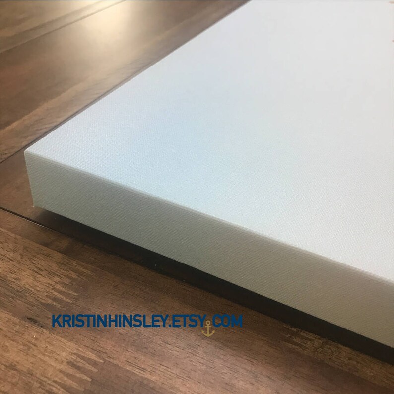 May include: A close-up of the edge of a light blue canvas print with a black frame. The print is on a wooden surface.