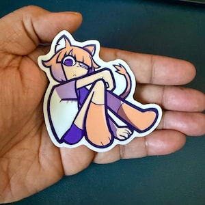 May include: A die-cut vinyl sticker featuring a cartoon character with peach-colored skin, cat ears, and a tail. The character has purple eyes, wears a white and purple shirt, and is depicted in a seated, hunched pose with a thick dark purple outline.