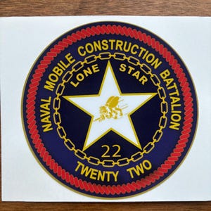 May include: A blue and gold circular sticker with the text "NAVAL MOBILE CONSTRUCTION BATTALION LONE STAR 22 TWENTY TWO" in a gold chain-link border. The sticker has a red rope border.