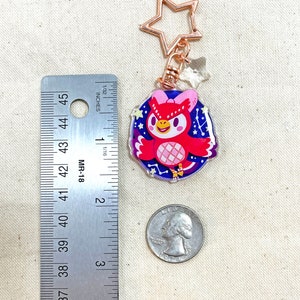 K&ouml;nnte beinhalten: Ein goldener Schl&uuml;sselanh&auml;nger mit einem sternf&ouml;rmigen Ring, an dem ein roter Eulenanh&auml;nger mit einer rosa Schleife und einem Sternenmuster auf den Fl&uuml;geln h&auml;ngt. Der Hintergrund ist eine blaue und violette Galaxie.