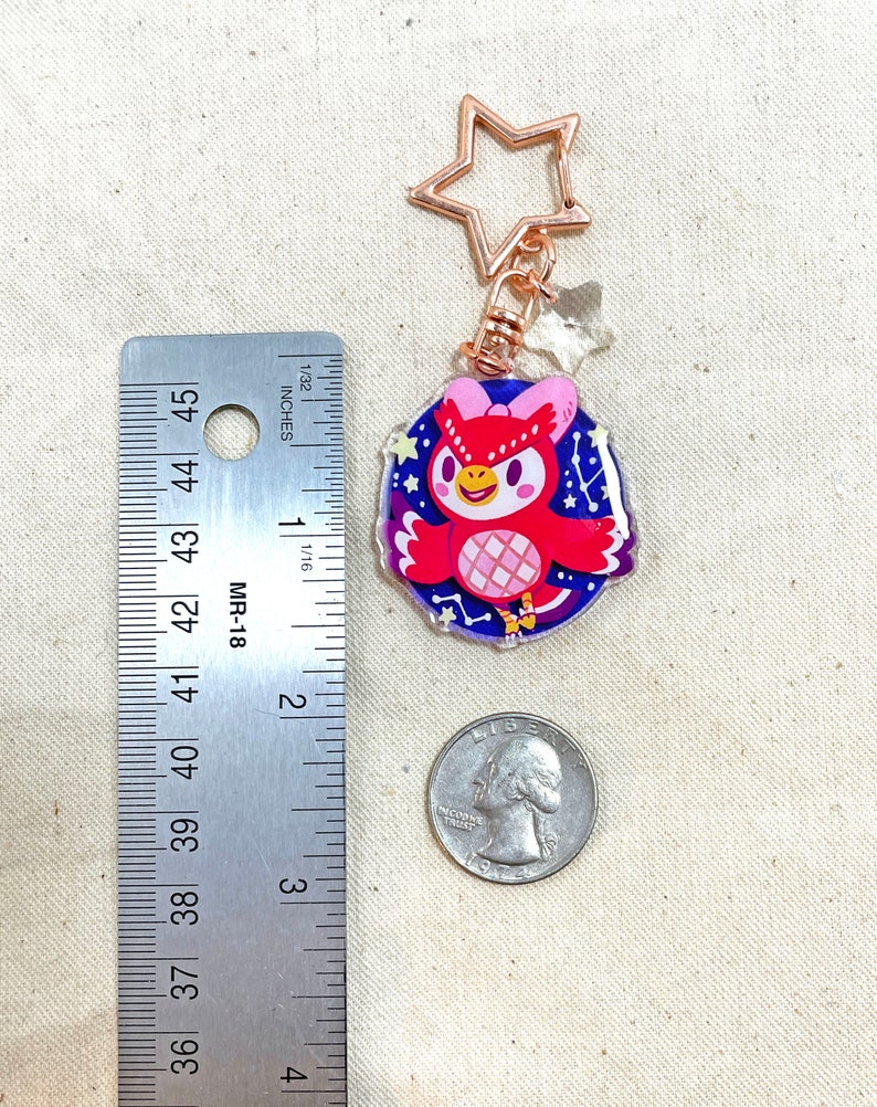K&ouml;nnte beinhalten: Ein goldener Schl&uuml;sselanh&auml;nger mit einem sternf&ouml;rmigen Ring, an dem ein roter Eulenanh&auml;nger mit einer rosa Schleife und einem Sternenmuster auf den Fl&uuml;geln h&auml;ngt. Der Hintergrund ist eine blaue und violette Galaxie.