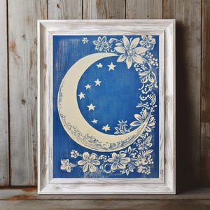 Puede incluir: Impresión enmarcada con una luna creciente y estrellas crema sobre fondo azul. Diseños florales y de hojas blancas rodean la luna. El marco es blanco con acabado envejecido. Obra de arte decorativa de temática celestial.