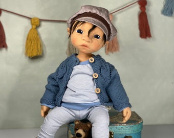 Puede incluir: Una muñeca con un cárdigan azul de punto, una camisa a rayas y mocasines marrones. La muñeca está sentada en una maleta azul con un diseño de mapa del mundo. Un oso de peluche marrón está sentado junto a la muñeca.