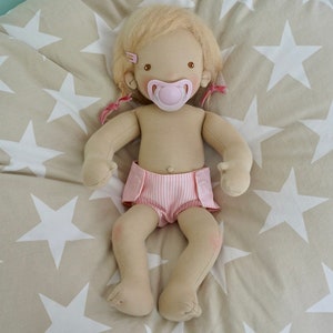 May include: A handmade cloth doll with blonde hair wearing a pink and white striped diaper and a pink pacifier. The doll is lying on a beige fabric with white stars.