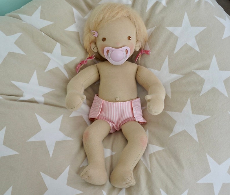 May include: A handmade cloth doll with blonde hair wearing a pink and white striped diaper and a pink pacifier. The doll is lying on a beige fabric with white stars.
