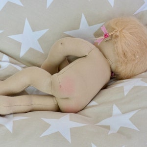 May include: A handmade cloth doll with blonde hair and a pink bow, lying on a beige and white star patterned fabric.