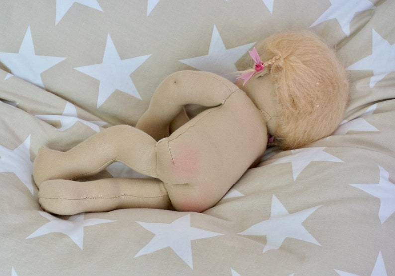 May include: A handmade cloth doll with blonde hair and a pink bow, lying on a beige and white star patterned fabric.