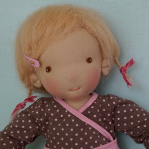 May include: A handmade doll with blonde hair, brown eyes, and rosy cheeks. The doll is wearing a brown and white polka dot dress with a pink trim.