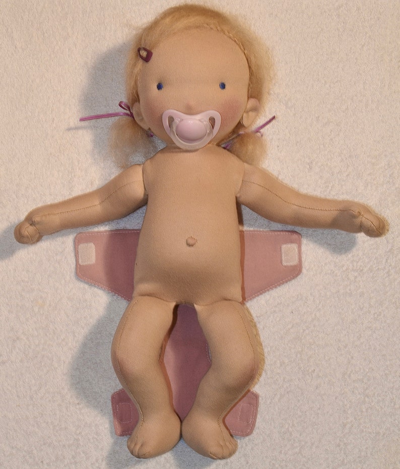 May include: A doll with blonde hair wearing a pink diaper and a pacifier. The doll has blue eyes and a light brown complexion.
