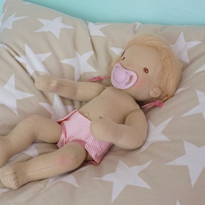 May include: A handmade cloth doll with blonde hair, wearing a pink and white striped diaper, lying on a beige pillow with white stars.