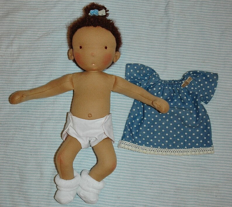May include: A brown-skinned doll with brown hair wearing a white diaper and white booties. The doll is holding a blue and white polka dot dress with white lace trim.