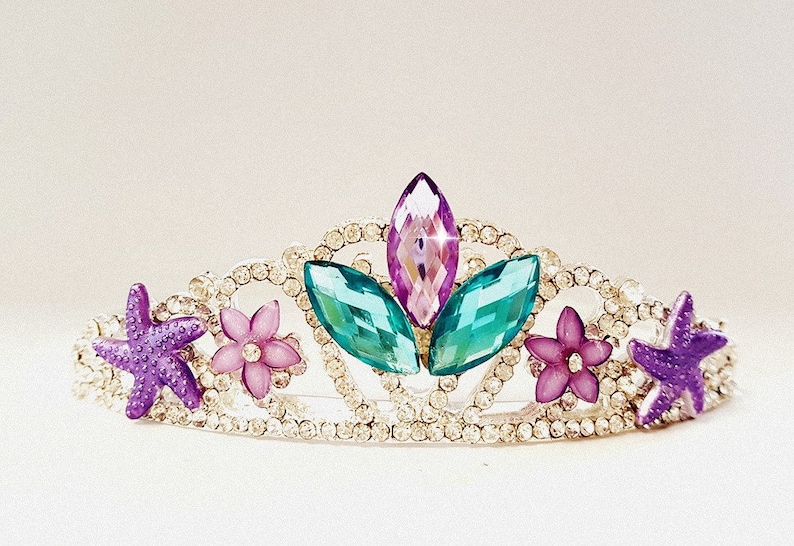 Puede incluir: Una tiara plateada con pedrer&iacute;a transparente, con una gema central morada y rosa, flanqueada por gemas turquesas y adornada con estrellas de mar moradas y flores rosas. Un adorno para la cabeza decorativo.