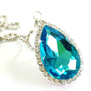 Puede incluir: Un collar con colgante en forma de l&aacute;grima con una gran gema facetada de color turquesa. La gema est&aacute; rodeada por un halo de peque&ntilde;os cristales transparentes engastados en un metal plateado. El collar tiene una delicada cadena plateada.