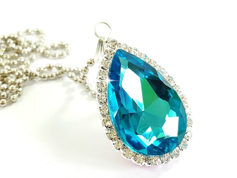 Puede incluir: Un collar con colgante en forma de l&aacute;grima con una gran gema facetada de color turquesa. La gema est&aacute; rodeada por un halo de peque&ntilde;os cristales transparentes engastados en un metal plateado. El collar tiene una delicada cadena plateada.