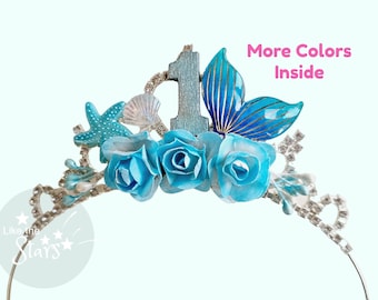 May include: A mermaid-themed tiara with a silver band, adorned with turquoise roses, a starfish, and a seashell. The tiara features a wooden number 1 and a blue mermaid tail. Text on the image says "More Colors Inside".
