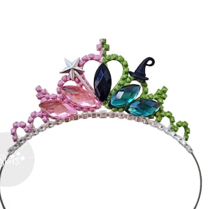 May include: A split-coloured tiara featuring a pink side with a silver star wand charm and a green side with a small black witch hat charm. The centre is adorned with a black marquise-cut gem, flanked by pink and teal marquise-cut crystals.