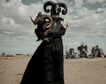 May include: A person wearing a black dress and a black horned mask holds a brown and black ram in their arms. The person is standing in a desert landscape with a cloudy sky.
