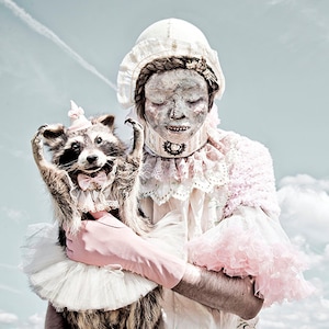 Puede incluir: Una persona con un vestido blanco con volantes rosas y un gorro blanco sostiene un mapache que lleva un tutú blanco y un lazo rosa. El mapache lleva una pequeña corona en la cabeza.