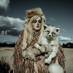 Op de afbeelding: Een vrouw met een bontmuts en een bontjas gemaakt van gescheurd papier houdt een witte vos in haar armen. De vrouw heeft wit haar en draagt ​​zware make-up. De achtergrond is een bewolkte lucht en een zanderig landschap.