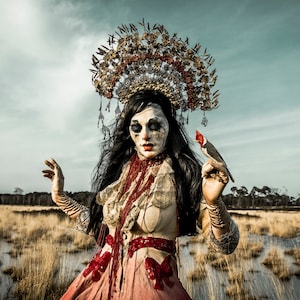 May include: A woman wearing a large, ornate headdress with a red cardinal perched on her finger. She is wearing a white and red dress with a lace bodice. The background is a blurred landscape.