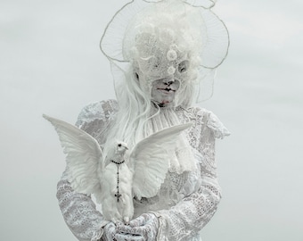May include: A woman in a white lace dress and a white veil holds a white dove with a silver cross around its neck. The dove is perched on her hand.