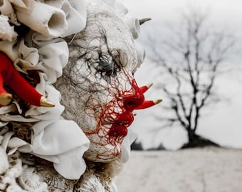 May include: A person wearing a white, lacey costume with red string details and a white face mask. The person's face is covered in red string and they have a red, string-like mouth.