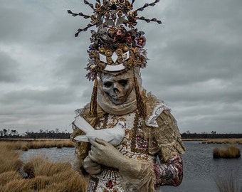 May include: A person wearing a white and gold costume with a skull face mask and a large, ornate headpiece. They are holding a white dove in their hands. The background is a blurry image of a body of water and reeds.