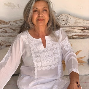 May include: A woman with gray hair is wearing a white embroidered tunic top with a V-neck. She is sitting on a white bench with a white pillow behind her.