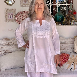 May include: White cotton tunic top with intricate embroidery in a floral pattern. The top has a V-neckline and long sleeves. The model is wearing white pants.
