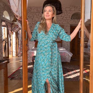 May include: A woman wearing a long, teal blue and white patterned wrap dress with a side slit. The dress has a V-neckline and 3/4 sleeves. She is standing in a doorway with a white brick wall behind her.