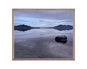 May include: A photograph of a large, dark rock in a shallow body of water. The water reflects the sky and mountains in the distance. The sky is a pale blue with white clouds.