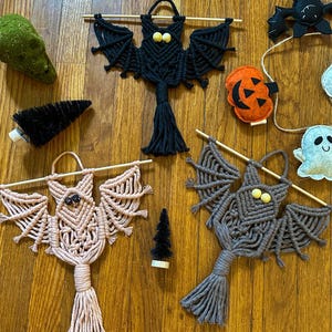May include: Three macrame bat wall hangings in black, light pink, and grey, each featuring wooden bead eyes and a tassel. The collection includes a felt pumpkin, a felt ghost, a black felt bat, and two small black bottle brush trees.