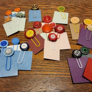 May include: A collection of colorful paper clips with various buttons attached. The buttons are in different colors, shapes, and sizes. The paper clips are attached to small pieces of colored paper.