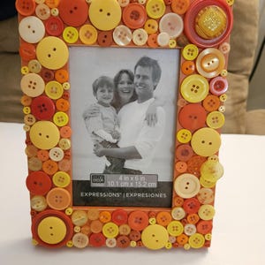 May include: A rectangular photo frame decorated with various sizes and shades of orange, yellow, and white buttons. The frame holds a black and white photo of a family. The frame is 4 in x 6 in (10.1 cm x 15.2 cm).