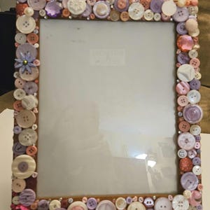 May include: A rectangular photo frame decorated with an assortment of pastel-colored buttons. The buttons are in shades of pink, purple, and white, and are arranged around the frame's border. The frame has a brown border and a glass insert.
