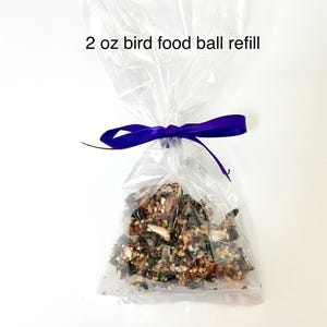 May include: A clear plastic bag filled with bird food, tied with a purple ribbon. The bag contains a 2 oz bird food ball refill. The bird food appears to be a mix of seeds, nuts, and other ingredients.