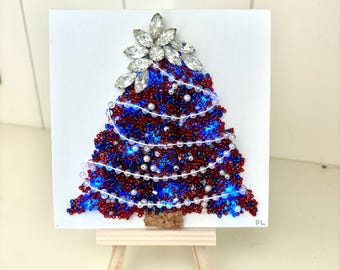 May include: A handcrafted Christmas tree artwork on a small easel. The tree is made of red and blue beads, adorned with white pearl-like beads and a sparkling silver starburst at the top. The base is a cork piece.
