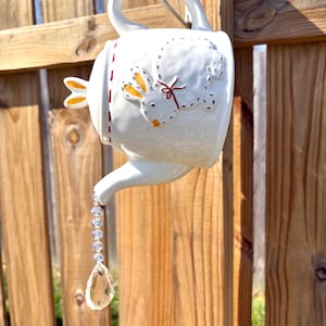 May include: White ceramic teapot shaped like a bunny with red stitching detail and a crystal teardrop hanging from the spout.
