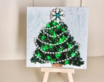 May include: A small, square Christmas tree artwork. The tree is made of green beads with white bead garland and a star-shaped topper. The artwork is mounted on a small wooden easel.