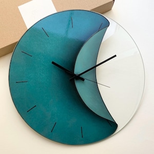 Può includere: Un orologio in vetro verde acqua e bianco con lancette nere. Il quadrante dell'orologio è diviso in due sezioni, una verde acqua e una bianca, con una linea curva che separa le due sezioni.