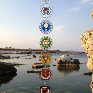 Set of 7 chakra suncatchers hanging vertically outdoors by the sea. Handmade wall hanging chakras decor with mandala glass details, spiritual gift for meditation lovers, yoga spaces and mindfulness inspired homes.