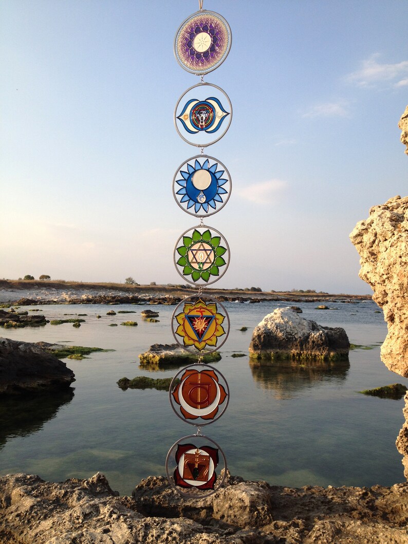 Set of 7 chakra suncatchers hanging vertically outdoors by the sea. Handmade wall hanging chakras decor with mandala glass details, spiritual gift for meditation lovers, yoga spaces and mindfulness inspired homes.