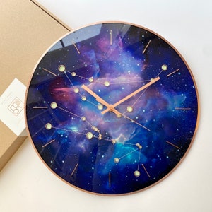 May include: A round clock with a dark blue background featuring a galaxy design with constellations and glowing stars. The clock hands are gold and the clock face is surrounded by a gold rim.