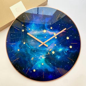 May include: Circular wall clock with a cosmic design. The clock face displays a blue and turquoise nebula pattern with constellations and gold-coloured hands. The clock has a copper-coloured rim.