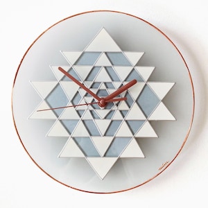 May include: A white and gray geometric clock with copper-colored hands and a copper-colored frame. The clock face is a stylized design of overlapping triangles.