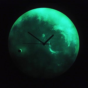 Puede incluir: Un reloj verde que brilla en la oscuridad con una esfera y agujas negras. La esfera del reloj es una imagen de un planeta con una pequeña luna.