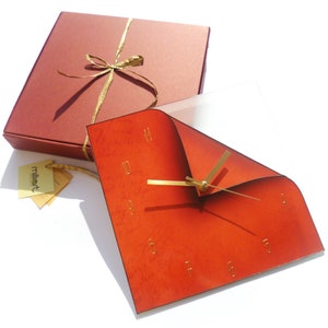 Puede incluir: Un reloj cuadrado rojo con agujas y números dorados. El reloj está en una caja con una cinta dorada. La caja es roja.