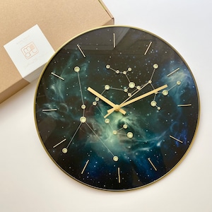 Puede incluir: Un reloj de pared redondo con marco de metal dorado y una esfera con un estampado de nebulosa espacial en tonos azul y verde. El reloj cuenta con manecillas doradas, marcadores de hora finos en dorado y puntos de constelación circulares claros.