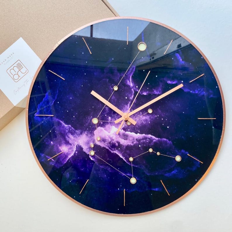May include: A round, purple and black galaxy-themed clock with gold hands and a rose gold rim. The clock face features a constellation of stars and a nebula.